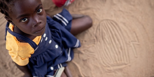 Bambina mozambicana ripresa dall alto è seduta a terra e guarda verso l alto. Indossa un vestito blu e una maglietta arancione.