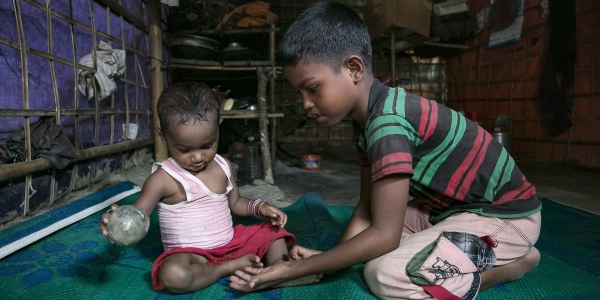 Un bambino di circa sei anni gioca seduto a terra su un tappeto verde con un bambino di circa un anno. Entrambi sono del Myanmar.