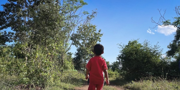un bambino di circa 5 anni di spalle vestito con maglietta a maniche corte e pantaloncini rossi cammina con infradito in una strada sterrata di un campo sfollati in myanmar