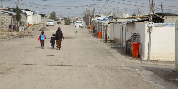 viale di un campo profughi con al centro in lontananza e di spalle tre persone che camminano
