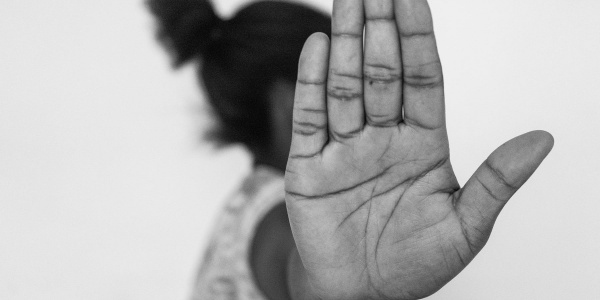 In primo piano la mano aperta in segno di stop di una ragazza nigeriana. Dietro la mano si intuisce un profilo di donna