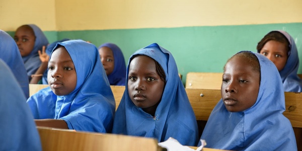 Tre bambine nigeriane della scuola primaria sono sedute a banchi scolastici in legno e guardano davanti a loro. Sono vestite con una divisa celeste e il loro capo è coperto.