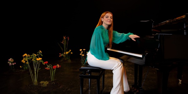 La cantante Noemi è seduta a un pianoforte su un set con alcuni fiori finti alle spalle. Indossa una camicia verde acqua e pantaloni bianchi.