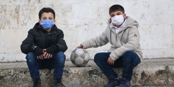 Due bambini siriani di 10 anni indossano la mascherina, un cappotto e dei jeans e sono seduti uno a fianco all altro su un gradino. Uno dei due tiene il braccio allungato lateralmente con la mano posata su un pallone, poggiato anch esso sul gradino.