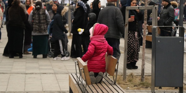 bambina con giacca rosa sola seduta su una panchina, sullo sfondo una folla di persone con trolley e zaini che fugge