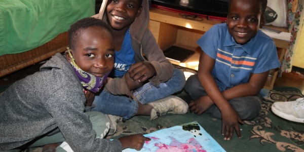 Tre bambini dello Zimbabwe sono seduti a terra intorno a un libro insieme. Hanno gli sguardi sorridenti e guardano in camera