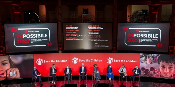 Palco ufficiale evento con locandine save the children e moderatori che discutono seduti sulle sedie