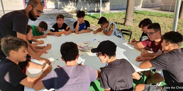 Studenti intorno a una tavola rotonda scrivono e studiano, un adulto li supervisiona