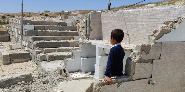 bambino palestinese di spalle con maglietta blu che guarda le macerie di una casa distrutta