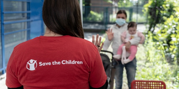 operatrice di save the children di spalle che saluta una mamma con un bimbo in braccio