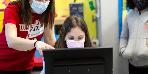 Una bambina davanti al pc e a fianco a lei un operatrice Save the Children accanto a lei indica lo schermo