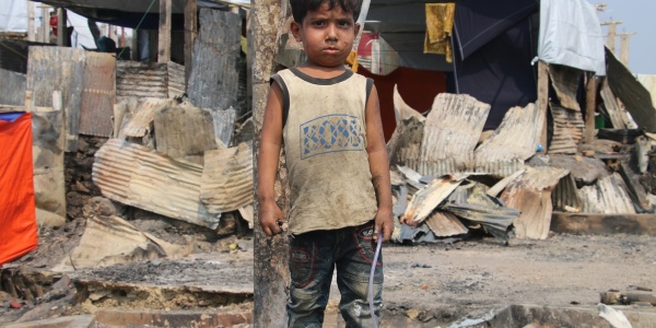 bimbo Rohingya in un campo profughi, vestito con canottiera e pantaloncini sporchi di terra, sullo sfondo un rifugio di lamiere fatiscente