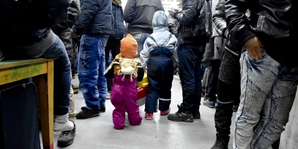 Due bambini molto piccoli di spalle sono in un ambiente chiuso molto affollato di cui si vedono soltanto le gambe e i piedi.