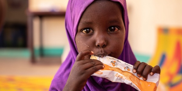 Bambina somala di pochi anni con chador viola in testa mangia una con le mani da una bustina di plumpynat
