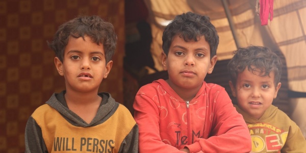 Tre bambini siriani seduti davanti alla tenda del campo in cui vivono guardano in camera. Il taglio è di mezzo busto