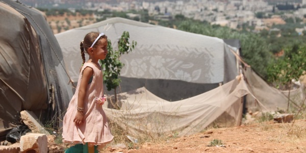 una bambina siriana di profilo cammina in un campo profughi. Dietro di lei si vede una tenda e un paesaggio arido. La bambina indossa un vestito rosa e i pantaloni di una tuta verde.