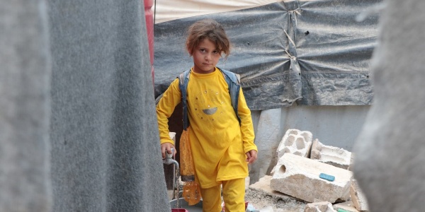 Una bambina vestita di giallo cammina in un campo profughi di cui si vedono alcune parti delle tende e dei rifiuti ingombranti dietro di lei.