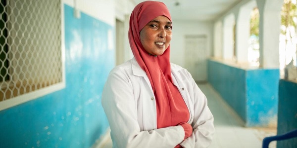 Operatrice sanitaria Fadumo che sorride con camice bianco e vero rosa attorno al volto con braccia incrociate mentre lavora nel nostro centro per la cura dei bambini malnutriti in Somalia