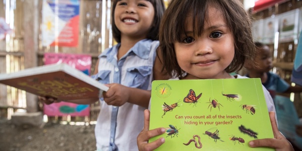 due bambine dentro un aula di scuola tengono in mano un libro a testa. Una di loro guarda in camera sorridente e laltra, sullo sfondo, ride.