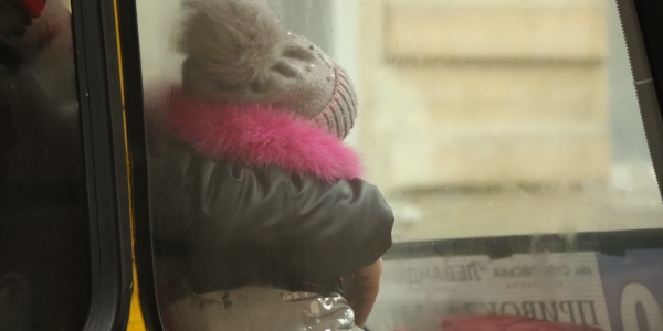 una bambina di spalle seduta su un autobus si vede dal finestrino. La bambina è vestita con un giubbotto pesante e un cappello di lana.