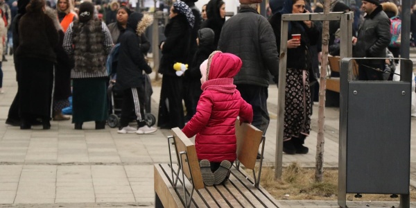 Una bambina con cappotto rosa di spalle è in ginocchio su una panchina. Davanti a lei una folla di persone.