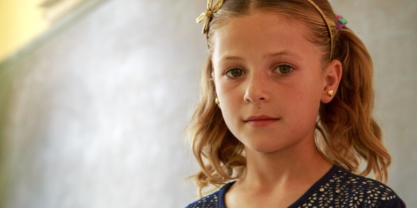 Un SMS per contrastare insieme la povertà educativa in Italia Bambina bianca bionda con i codini e un cerchietto rosa sui capelli sta davanti a una lavagna a scuola