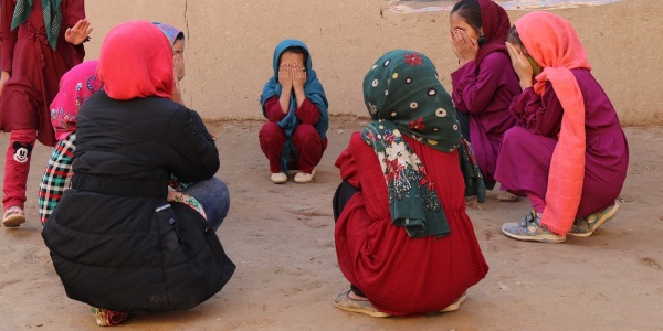bambine vestite in viole e rosso sono sedute in cerchio e si coprono il volto con le mani.