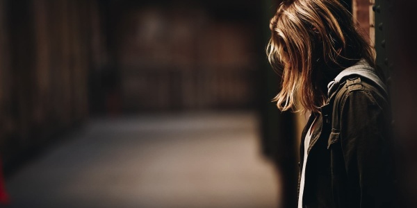 una ragazza bionda di profilo appoggiata con la schiena al muro. Guarda verso il basso e i capelli le coprono il viso. Indossa una giacca jeans e si vede sullo sfondo un corridoio vuoto e in semi oscurità.