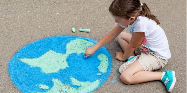 Una bambina bionda con la coda è seduta a terra sulla strada e disegna con i gessi un mappamondo. Lei è vestita con una maglietta a maniche corte bianca e dei pantaloncini marroni.