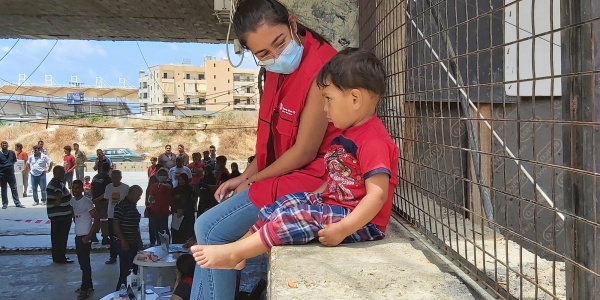 operatrice save the children con mascherina seduta su un muretto a fianco di un bimbo. Sullo sfondo varie presone che camminano. 