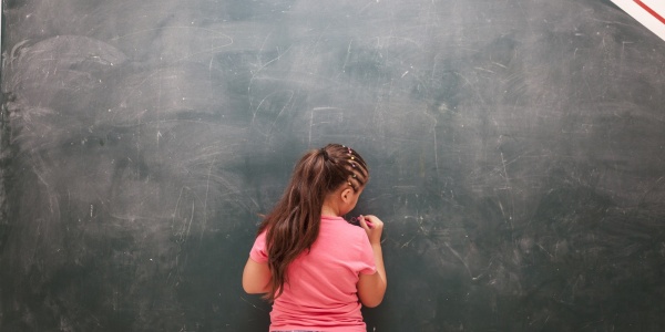 bambina di spalle davanti a una lavagna nera scrive con il gessetto