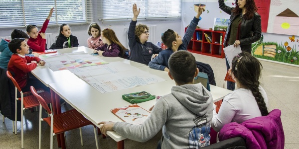 una decina di bambini seduti a un banco di scuola alzano la mano alla domanda della maestra